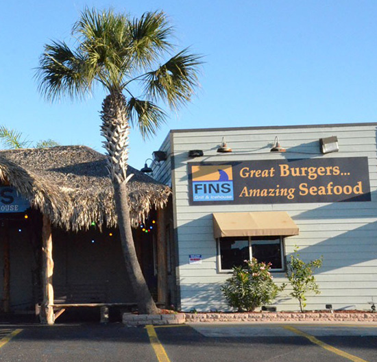 Fin’s Grill and Icehouse with a palm tree, thatched roof entrance, and seafood restaurant sign