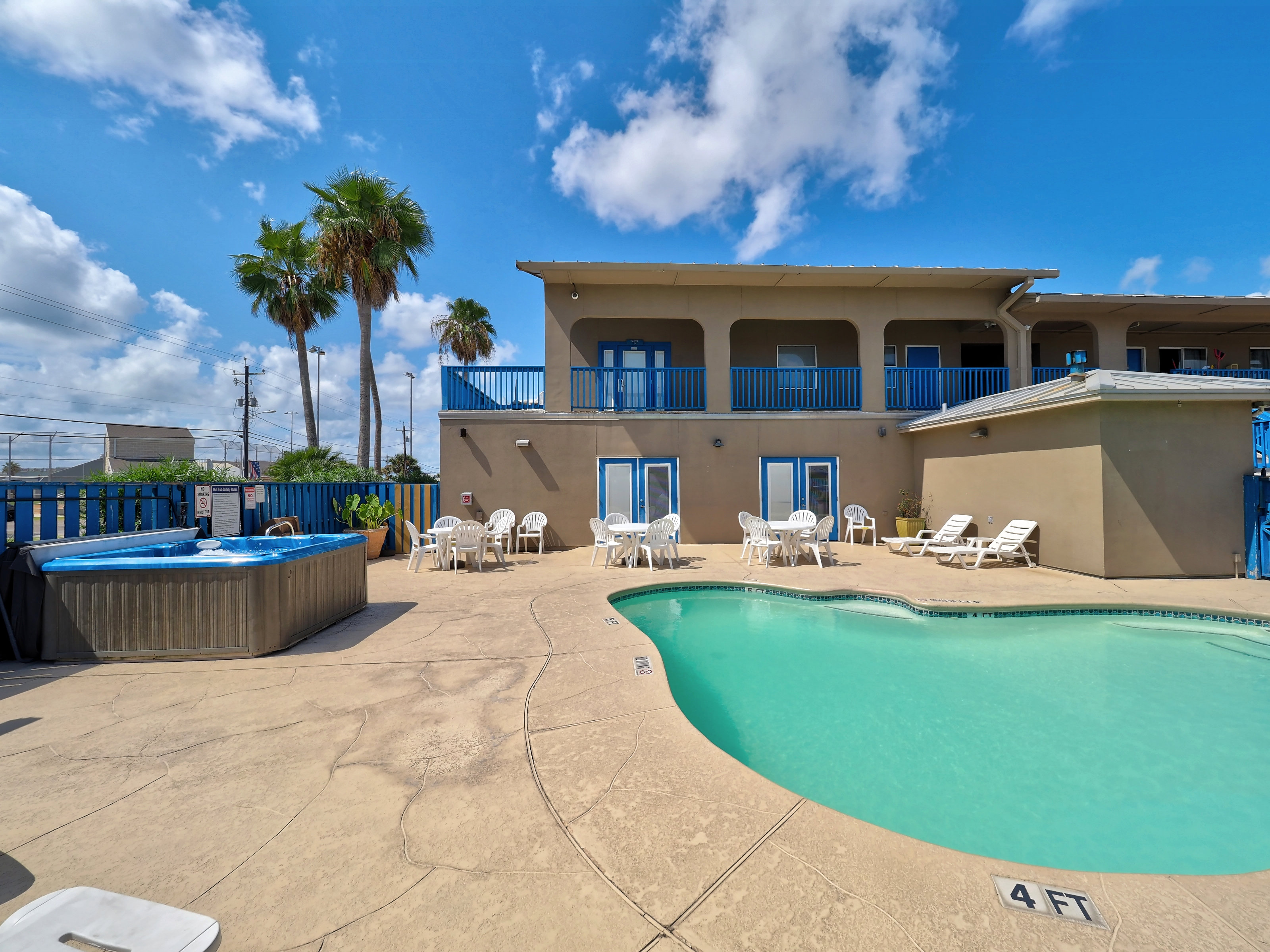 A hotel pool area with a pool, hot tub, lounge chairs, and palm trees under a cloudy sky.