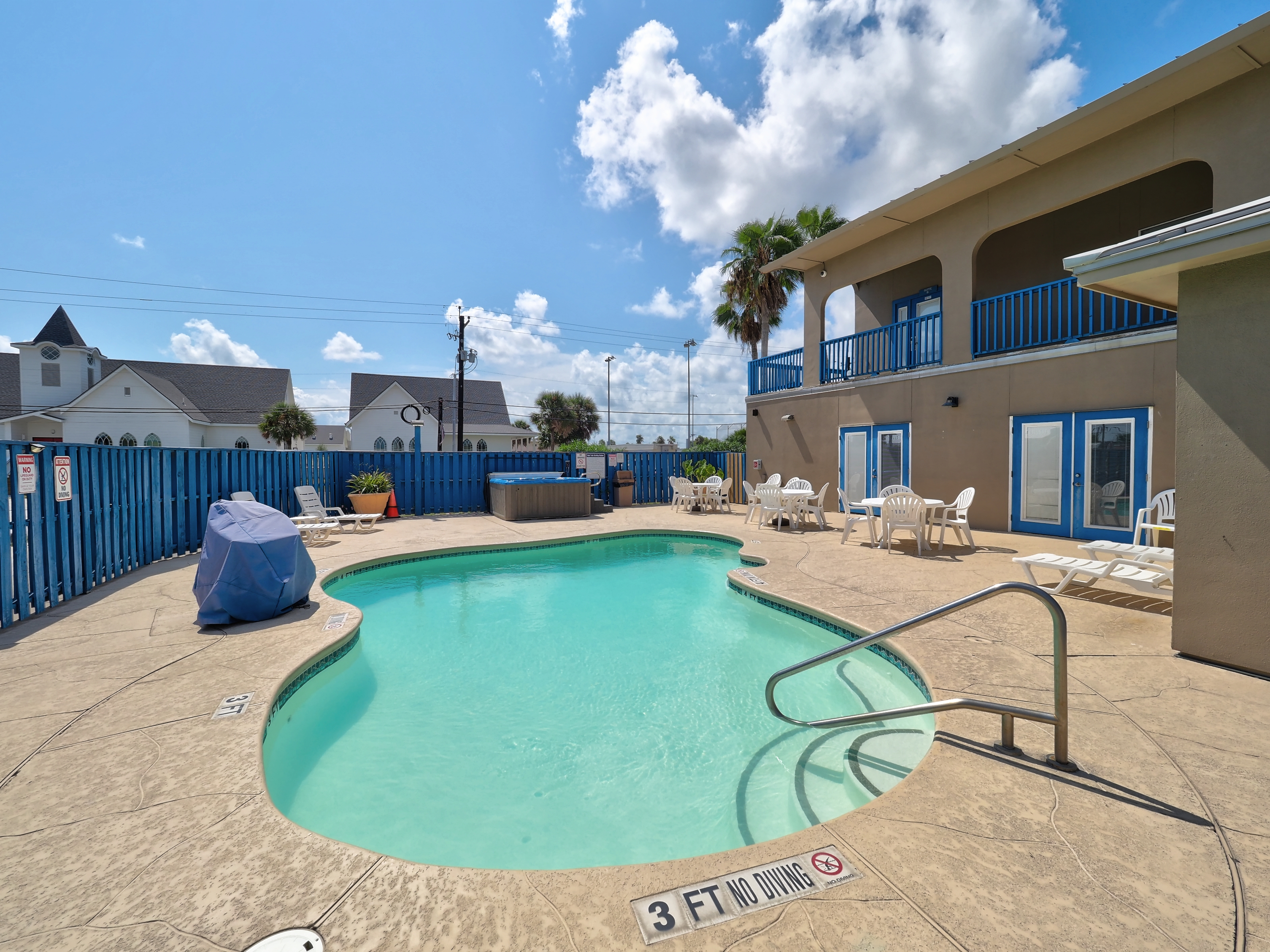 A kidney-shaped pool with a metal handrail, surrounded by white furniture and a blue fence, with a two-story building featuring blue-trimmed windows and balconies.