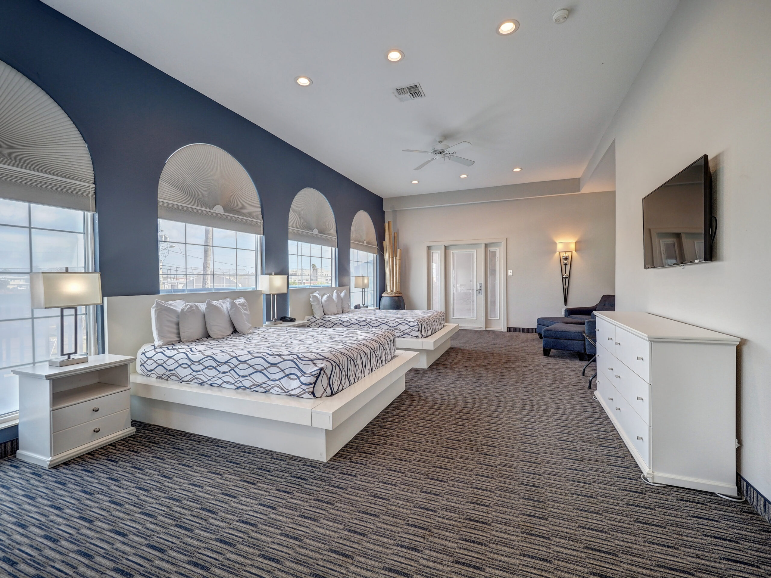 A spacious hotel room with two queen beds, a blue accent wall, arched windows, and a ceiling fan.