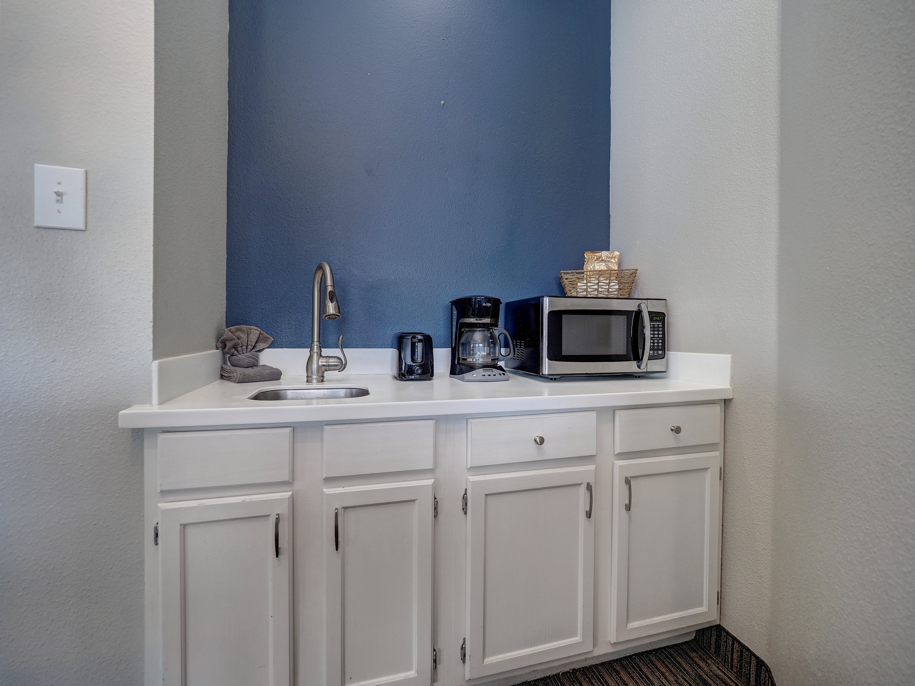 A small kitchenette with white cabinets, a microwave, coffee maker, and sink.